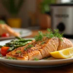 Perfectly cooked slow cooker salmon fillet garnished with fresh dill and lemon slices, served on a white plate with a side of roasted vegetables, set on a wooden countertop with a slow cooker in the background